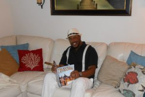 Wearing a black shirt, white pants and white suspenders, Antwan sits reading Travel & Leisure while holding a cigar in his right hand