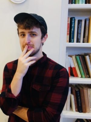 Antone, wearing flannel and a baseball cap, stands in front of a filled bookshelf